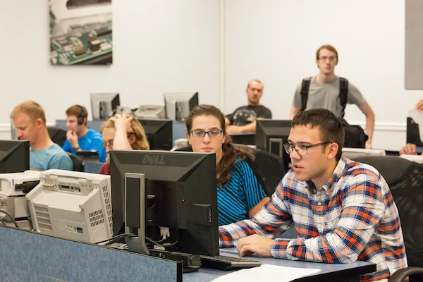 Students work at computers in a Valpo classroom, with others seated and standing in the background.