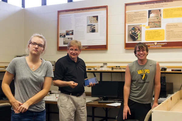 Three people stand in a Valpo classroom with engineering posters and computers in the background, highlighting the collaborative spirit fostered by Elite Partners.