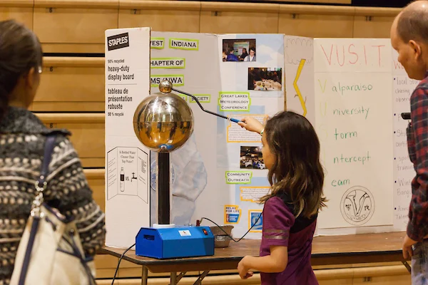 A girl touches a Van de Graaff generator at a STEM event, with adults watching and a display board behind her.