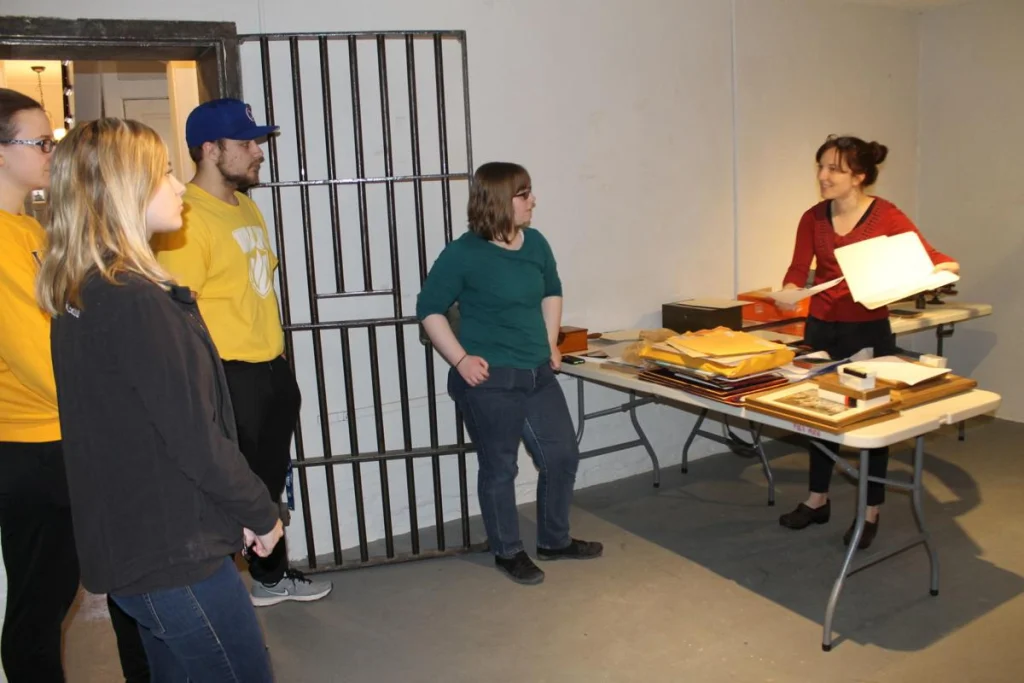 Four people from the History Club listen as a woman presents papers and artifacts on a table in a room with a barred door, sharing fascinating news about the past.