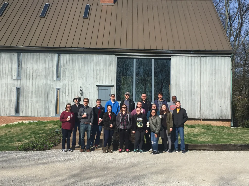 Sixteen people stand in a row outside a modern gray building on a sunny day, capturing a moment from their trip to New Harmony.