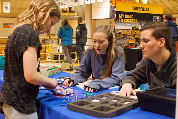 Three teens collaborate on a STEM electronics kit at a table during the STEM Faire, surrounded by informational booths and an atmosphere of innovation extraordinaire.
