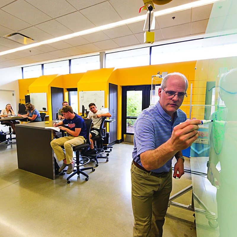 A teacher writes on a glass board while students watch and take notes in a bright Valpo classroom, inspiring future Elite Keen Partners.