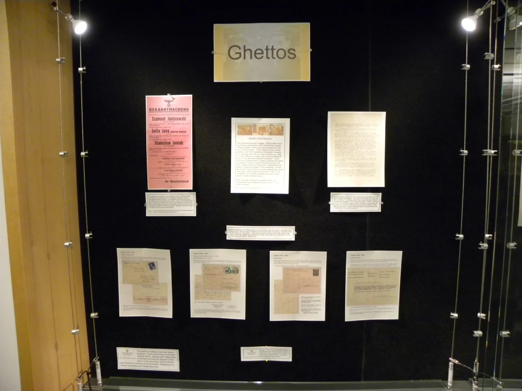A museum display case labeled "Ghettos" with various historical documents and letters on a black background, featured as part of the Holocaust Displays at the Christopher Center.