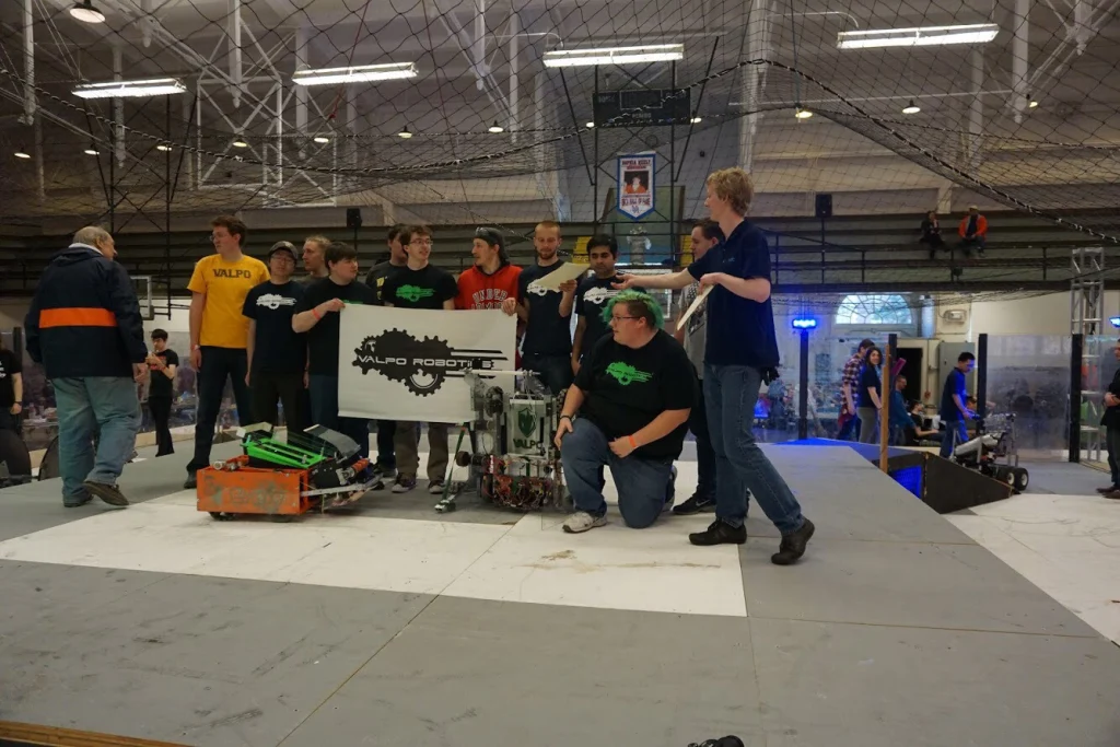 A group of teens pose with their robot and a sign reading "Valpo Robotics" at a design competition.