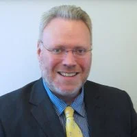 Smiling man with glasses in a suit, blue shirt, and yellow tie against a plain light background—Professor Ostoyich, Board Member of the Sino-Judaic Institute.