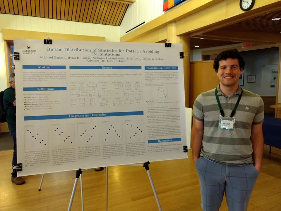 A man smiles, standing beside a large academic research poster on Permutation Patterns at an International Conference in an indoor Valpo setting.