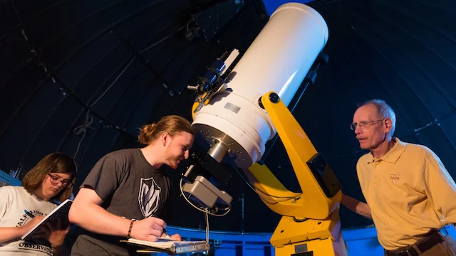 Two students take notes while observing the stars through a telescope, guided by an instructor in an observatory.