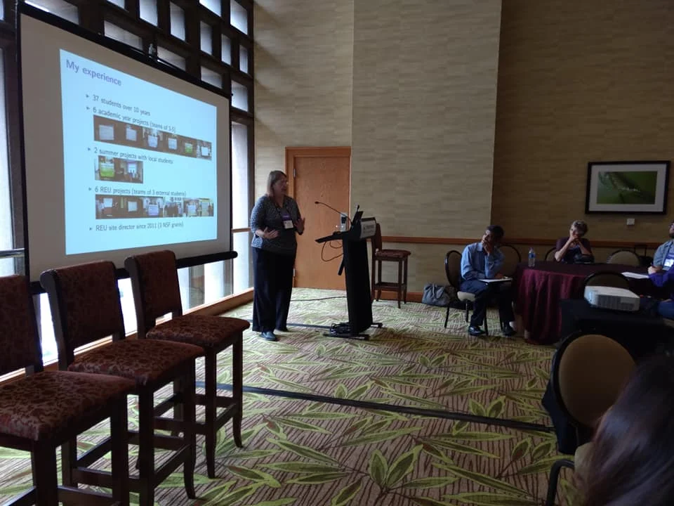 A woman presents slides to an audience in a conference room with large windows and empty chairs in front during Mathfest 2018.