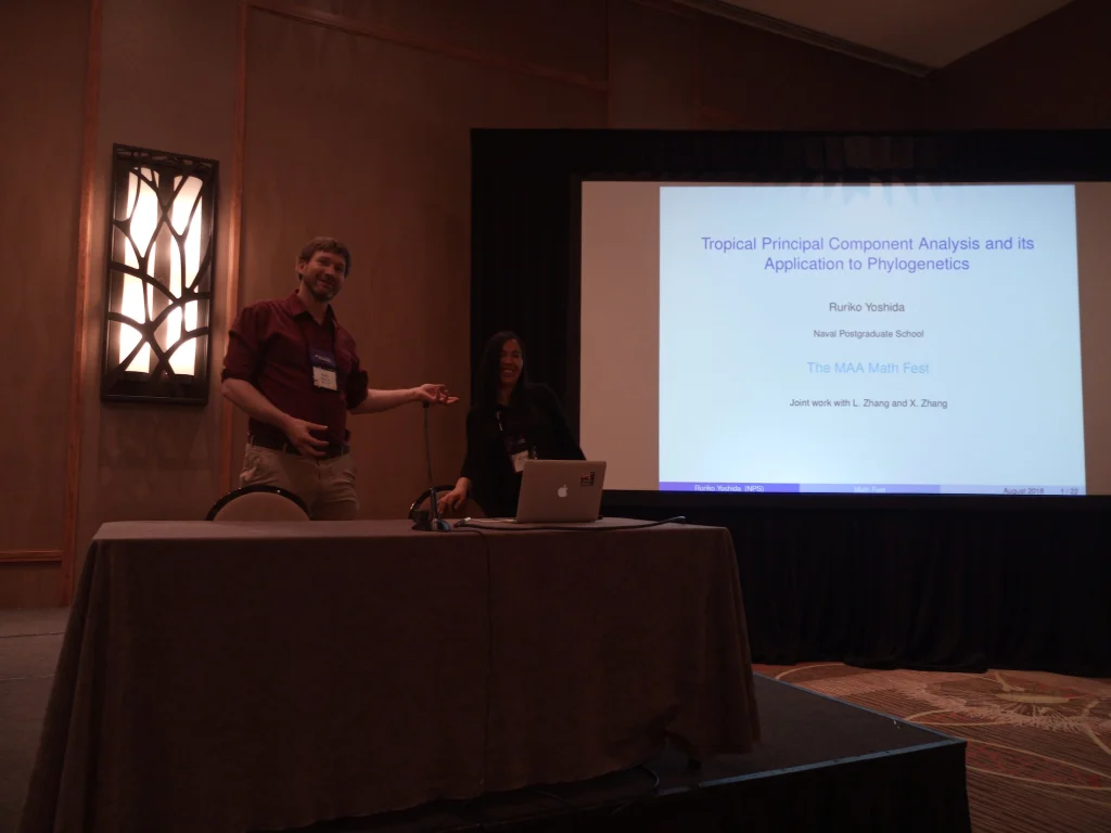 Two people stand by a podium near a screen displaying a math presentation in a dimly lit room, representing Valpo MST faculty at Mathfest 2018.