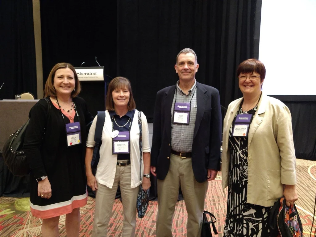 Four adults wearing conference badges—Valpo MST faculty—stand and smile together in a large room with a stage in the background at Mathfest 2018.