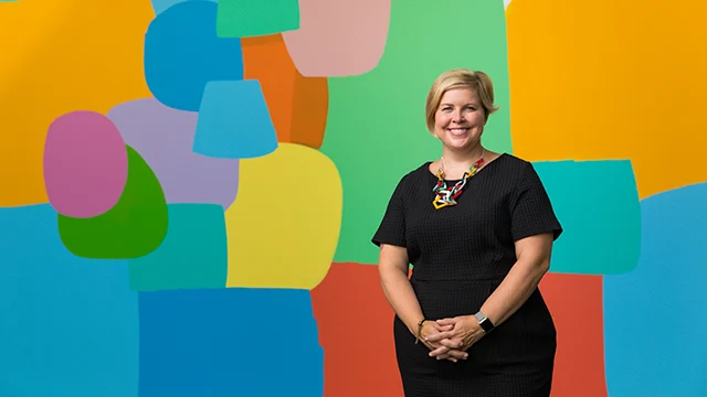 A smiling woman in a black dress stands in front of a colorful abstract mural with bold shapes, bridging past and present as she draws career inspiration from the dynamic art at the Brauer Museum.