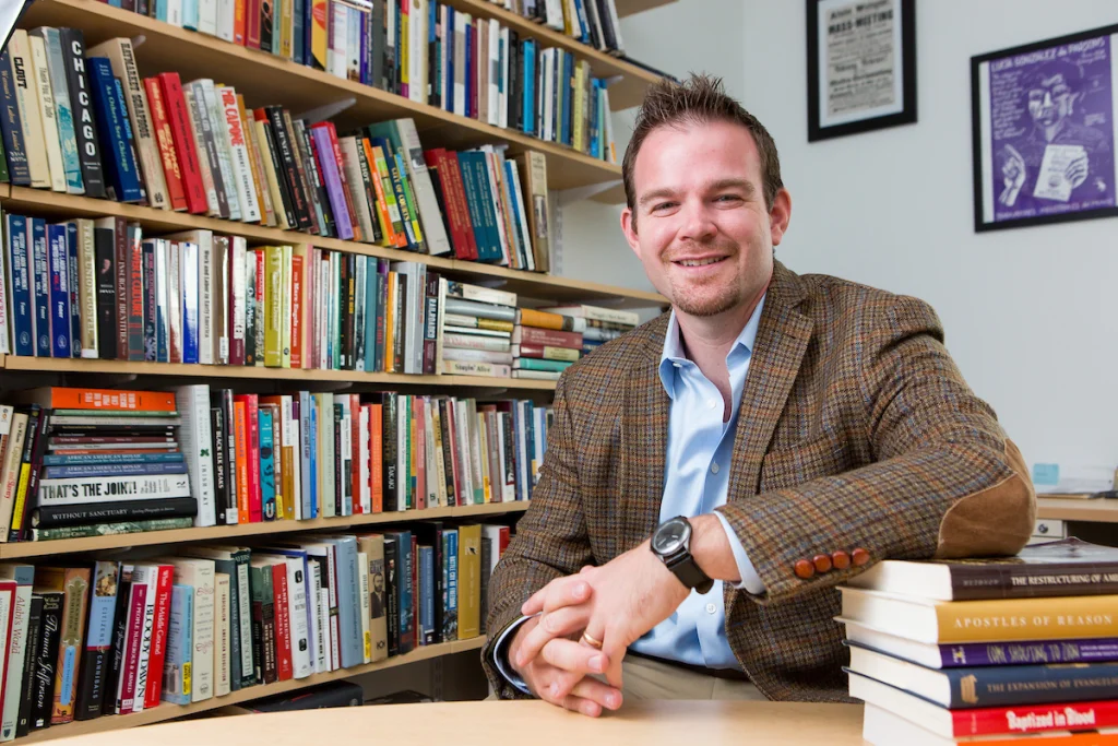 Man in a blazer sitting at a desk with bookshelves behind him, smiling at the camera—recipient of the Emerging Public Intellectual Award.