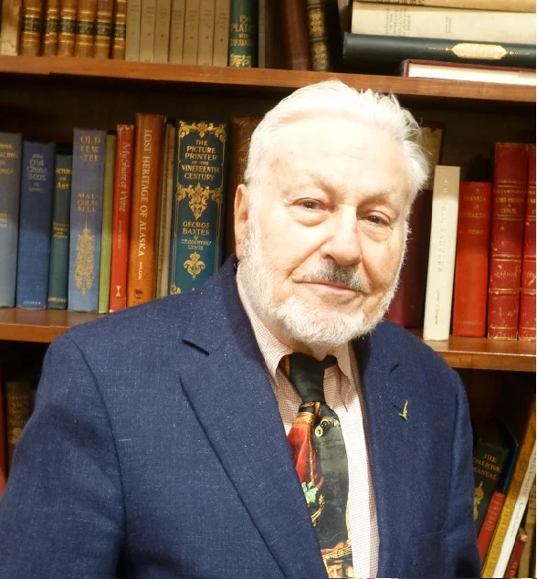 Older man with white hair and beard in a suit and tie, standing in front of a bookshelf filled with books, perhaps during a visit or event featuring Eric Kisch.