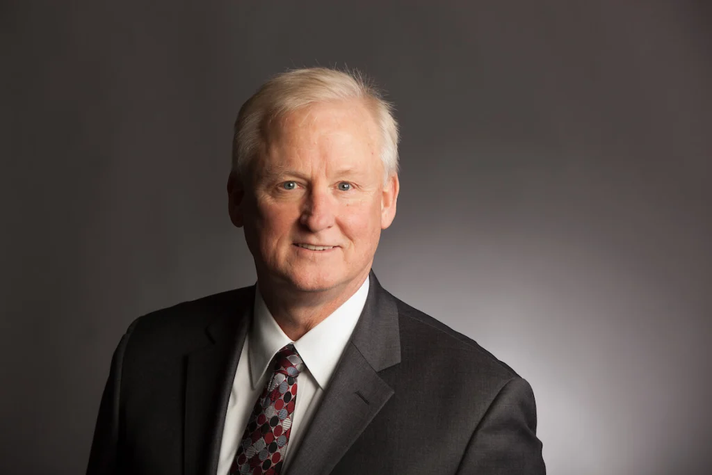 Man with short gray hair in a dark suit and patterned tie, smiling against a plain gray background; a distinguished member of the Valpo College of Business Review Committee.