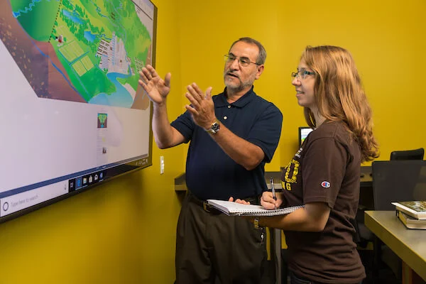 Two people discuss a colorful map on a screen; one gestures while the other takes notes on a notepad, exploring solutions in environmental engineering at Valpo.
