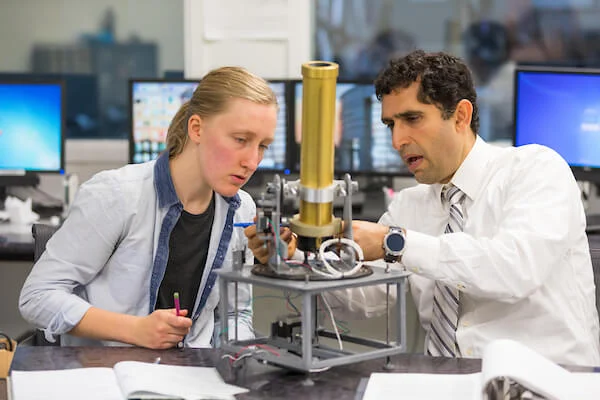 A Valpo Professor and a colleague collaborate on a scientific instrument in a lab filled with computers and equipment, enhancing the benefit of students through practical research experience.