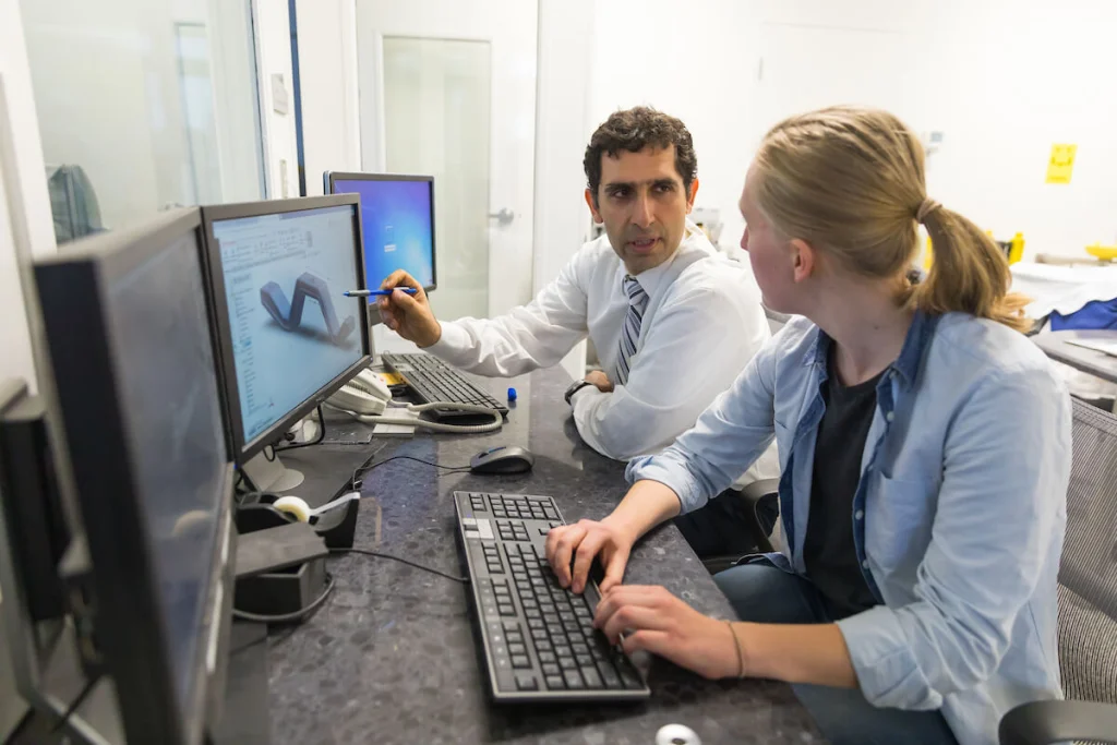A Valpo Professor in a shirt and tie discusses a 3D model on the computer screen with a woman at an office desk with two monitors, highlighting the benefit of students pursuing an additional degree.