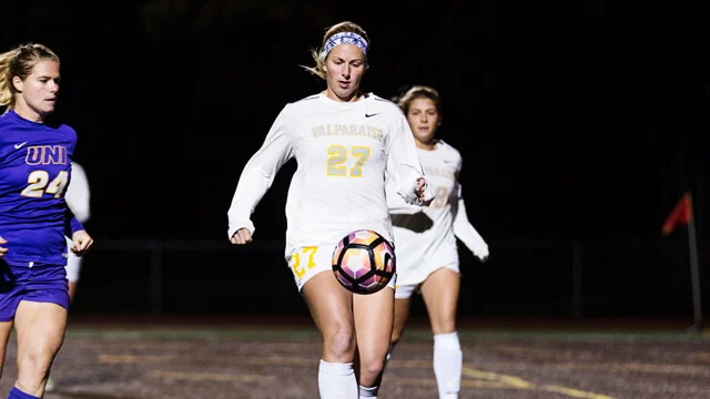 Soccer player in white jersey dribbles the ball at night, showcasing skills and determination—the same drive that leads to success on campus and a promising career, while opponents in purple run nearby.