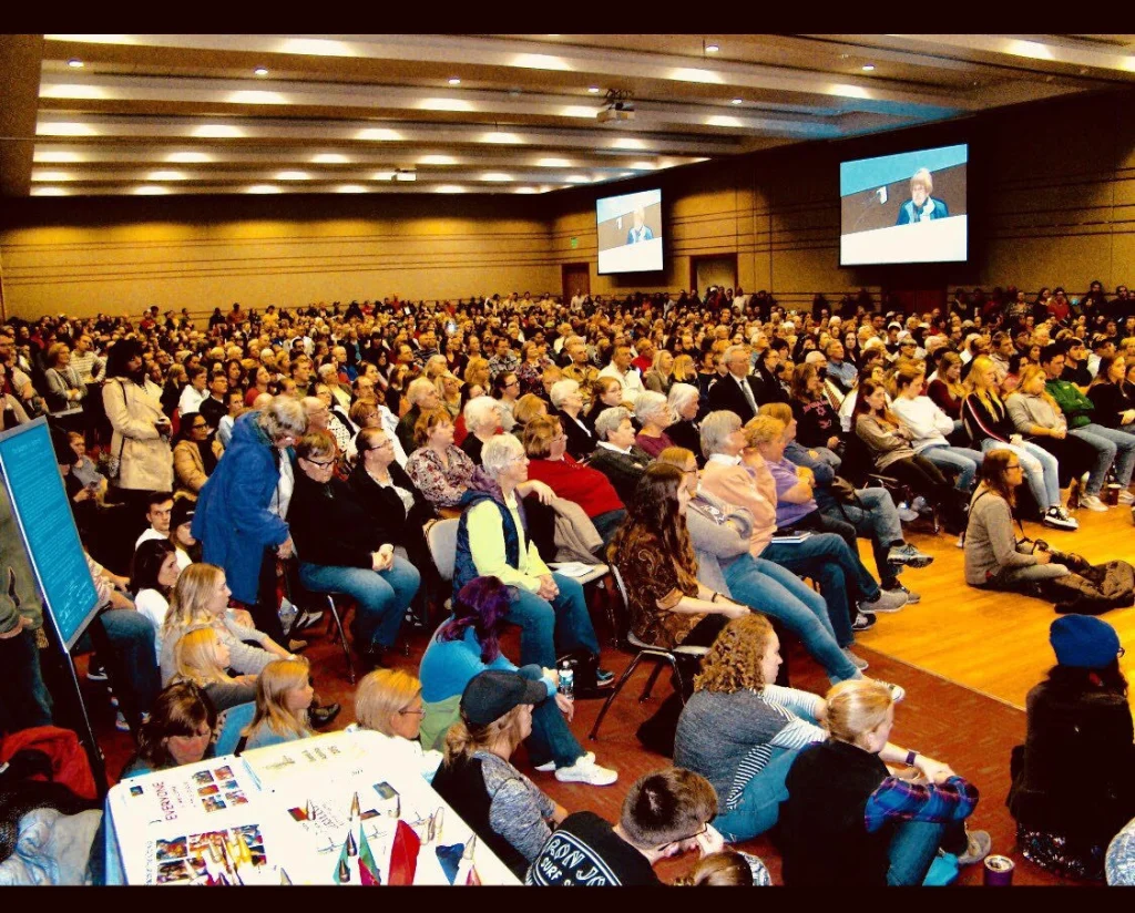 Large audience seated in a spacious hall, watching a speaker on stage with presentations on screens.