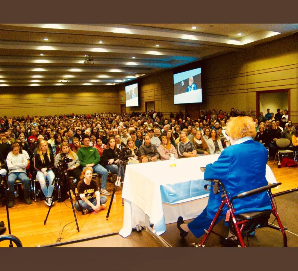 An elderly woman speaks to a large seated audience in a conference room with projection screens.