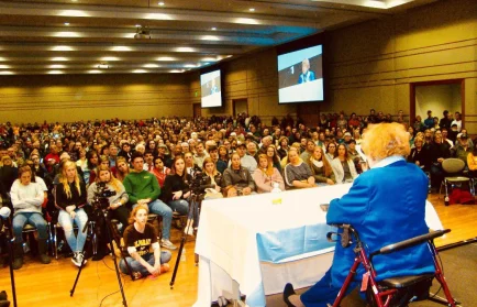 An elderly woman speaks to a large seated audience in a conference room with projection screens.