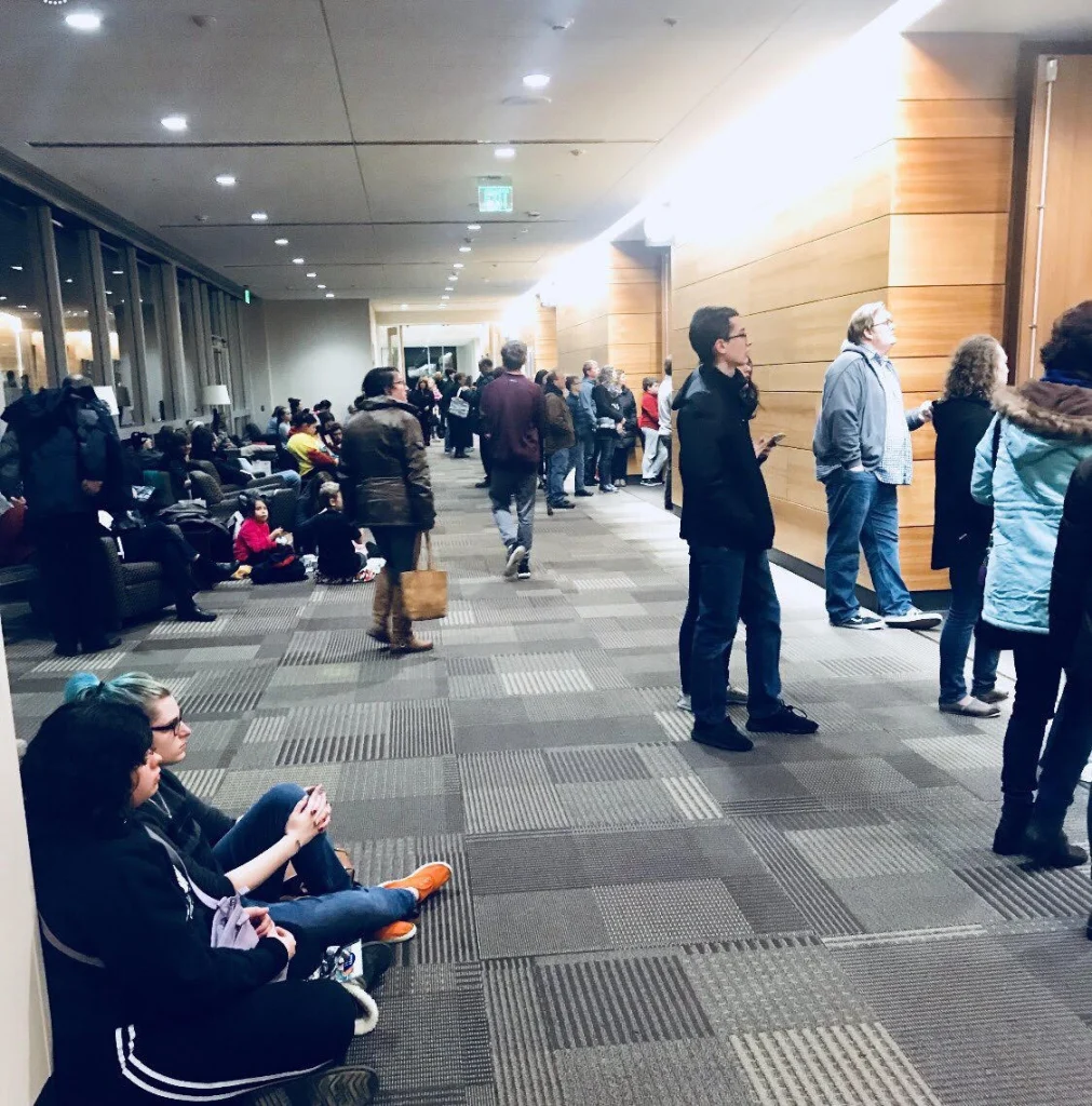 People wait in a long line and sit on the carpeted floor in a brightly lit hallway with wood-paneled walls.