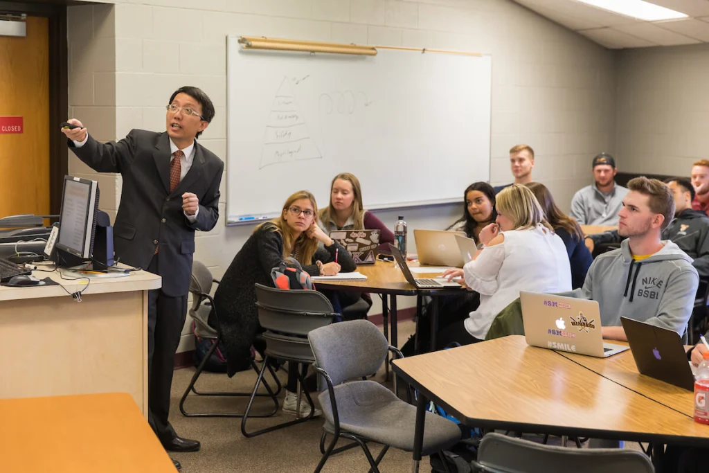 A New Faculty member gestures while teaching a group of students in a Finance Department classroom with laptops and notebooks.