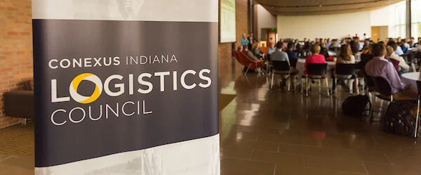 A banner for the Conexus Indiana Logistics Program stands near a room full of people seated at tables, highlighting the importance of supply chain innovation.