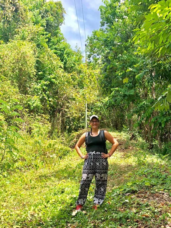 Hannah Purkey stands on a grassy trail surrounded by lush green trees, smiling and wearing patterned pants and a cap.