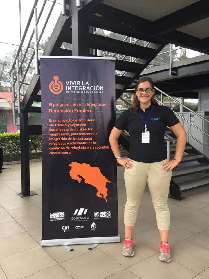 Smiling woman stands next to a “Vivir la Integración” employment program banner in an outdoor setting, celebrating success in a global environment.