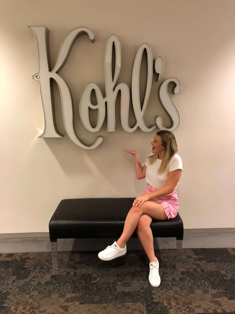 Woman sitting on a bench, smiling and pointing at a large "Kohl's" sign on the wall, showcasing her passion for careers in retail and business analytics programs.