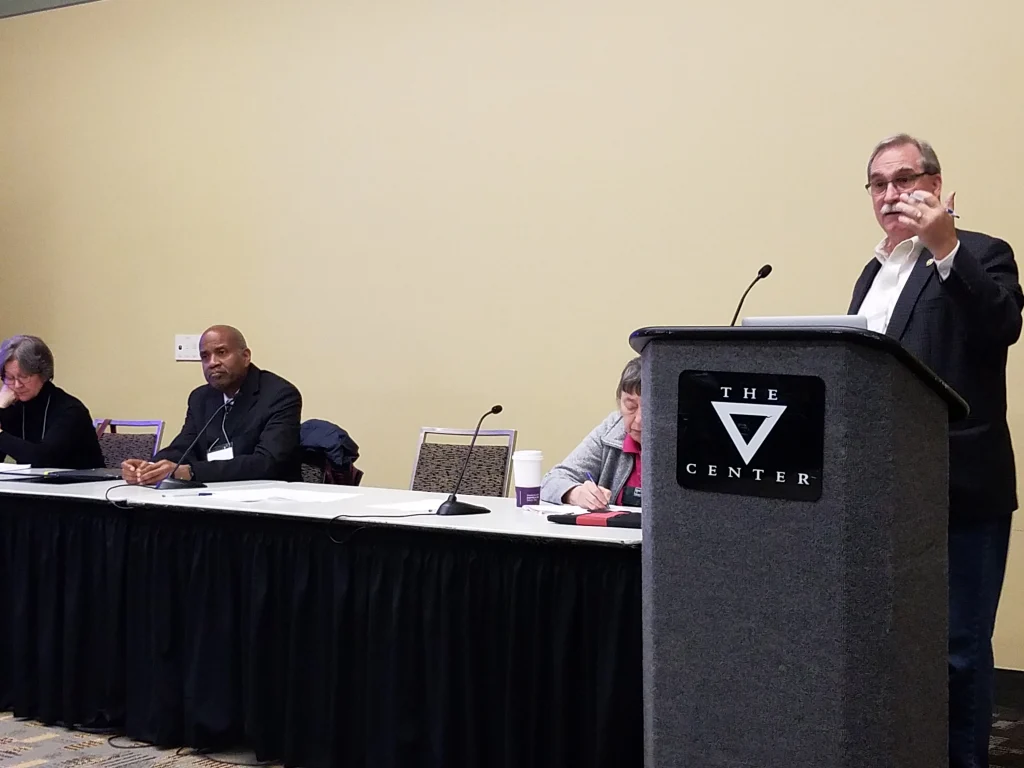 Four people sit at a panel table during the Joint Mathematics Meetings 2019, while one man speaks at a podium labeled "The V Center.