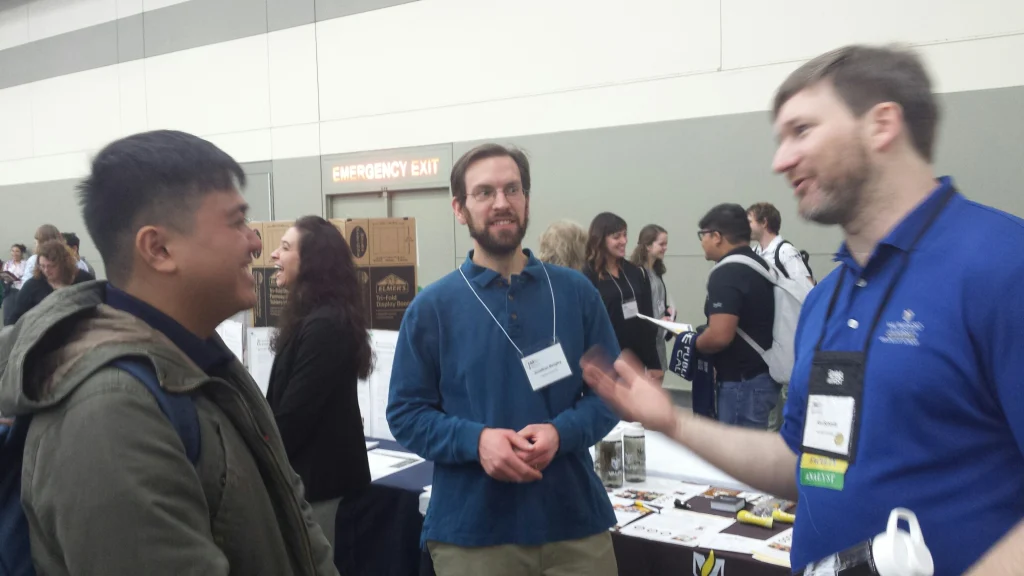 Three men talk and smile at a busy MST booth during the 2019 Joint Mathematics Meetings, with people and displays in the background.