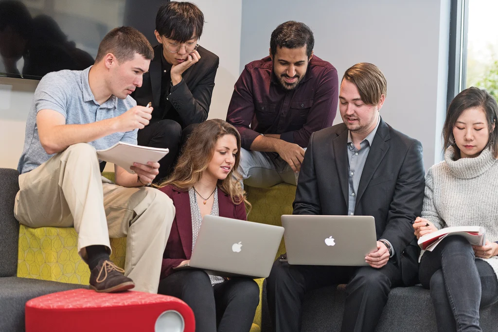 Six people sit and stand together, collaborating and working on laptops and notebooks in an office setting—an environment ideal for those in the Integrated Business and Engineering Bachelor’s Program.