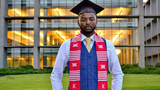 A graduate in a cap and gown poses outside Montel Hall at sunset, celebrating achievements both academically and athletically.