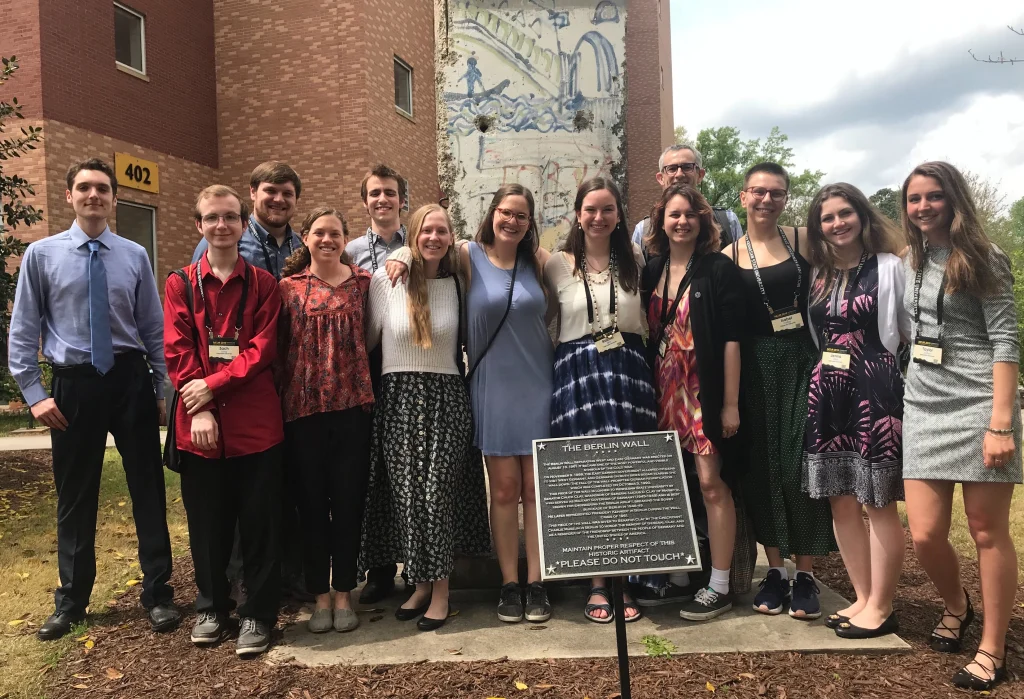 Thirteen CC Students pose and smile in front of a Berlin Wall monument and a historical plaque outdoors during the NCUR 2019 conference.