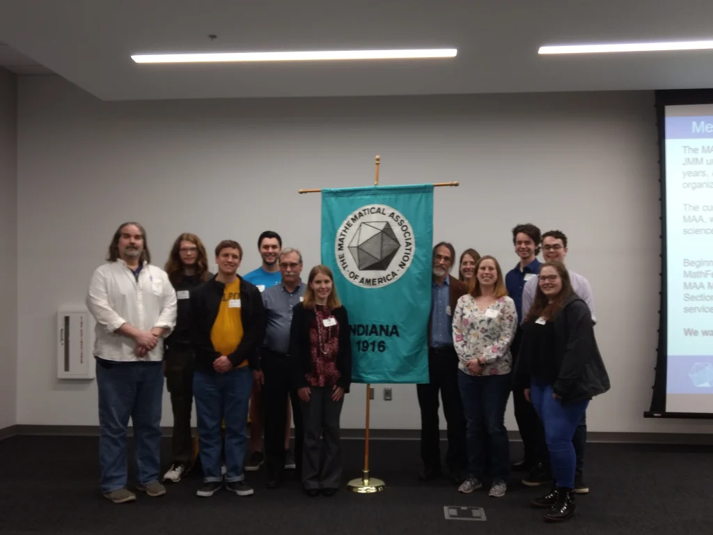 A group of twelve people stand by a Mathematical Association of America banner in a conference room at the Spring 2019 Indiana Section MAA meeting.