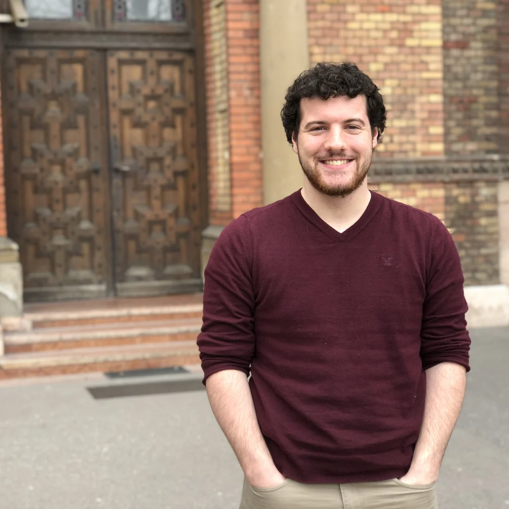 Man with curly hair and a maroon sweater smiles, standing outside near a brick building with wooden doors. Roth, a 2019-2020 Goldwater Scholar, radiates confidence in this candid moment.