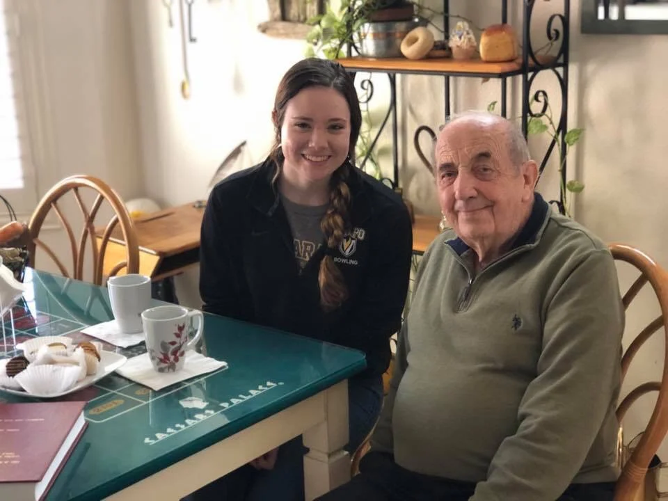 A young woman and Gary Sternberg, an elderly man, sit smiling at a kitchen table with coffee mugs and pastries, sharing stories about Dealer's Cards and the best moments in life.