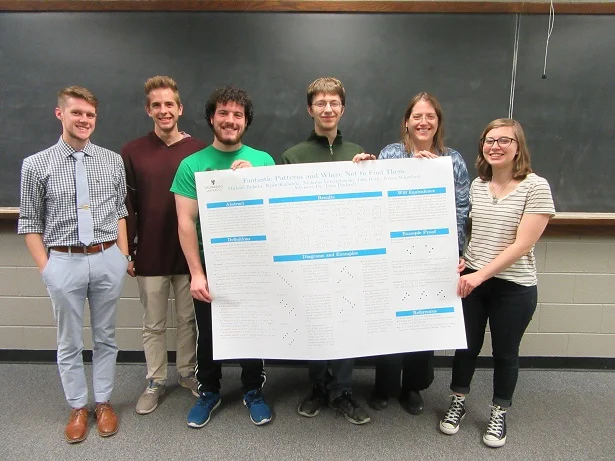 Six people stand and smile indoors, holding a large academic poster in front of a chalkboard, showcasing their collaborative publications from the MST program.
