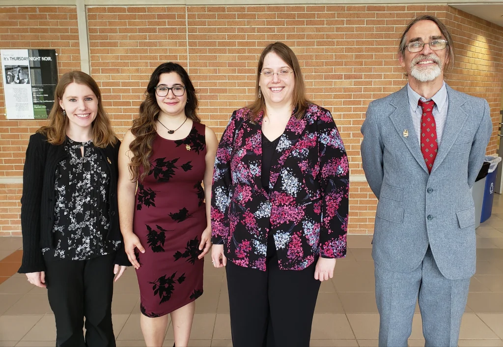 Four people stand indoors, smiling in business and semi-formal attire, with a brick wall behind them. Among them is Math major Tina Kalafatis, proudly wearing her Phi Beta Kappa pin.
