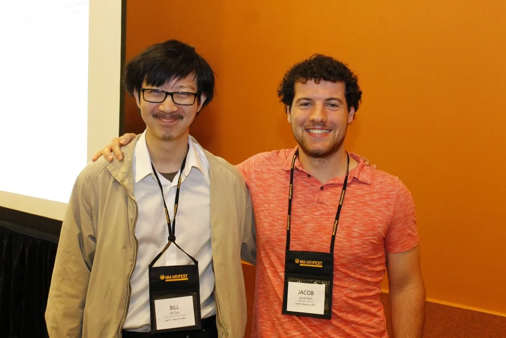 Two men with conference badges stand side by side, smiling, in front of an orange wall at Mathfest 2019.