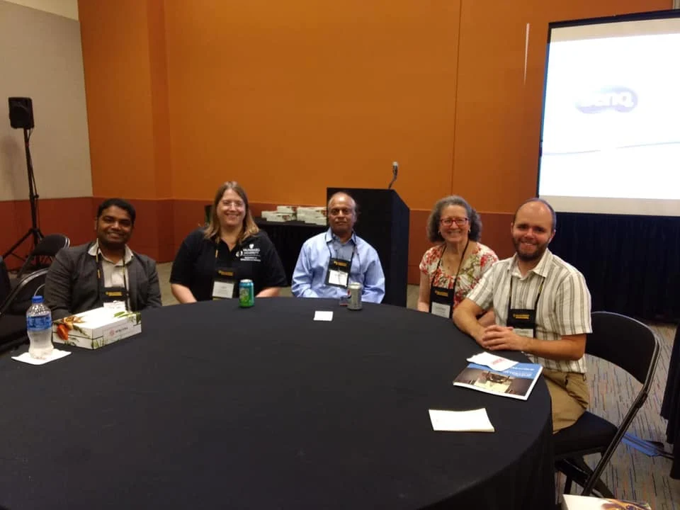 Five people sit around a round table in a conference room with a projector screen in the background, discussing MST topics at Mathfest 2019.
