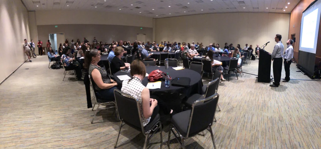 A large group of people seated at round tables listen to speakers presenting at the front of the Mathfest 2019 conference room.