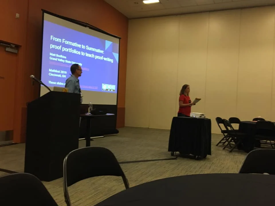 Two people are present in a conference room at Mathfest 2019, discussing a projected slide about teaching proof-writing.