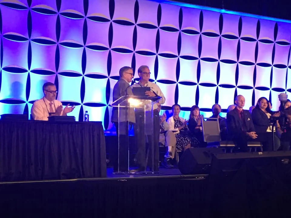 Two people stand at a podium on stage as others seated behind them applaud, with a blue-lit backdrop at Mathfest 2019, hosted by the MST.