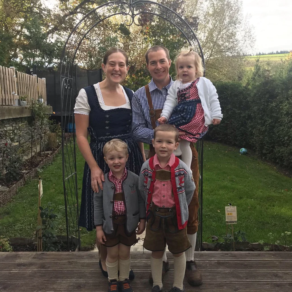 A family of five in traditional German attire stands smiling outdoors under a metal arch in a garden, perhaps celebrating Professor Venstrom's latest research in Germany.