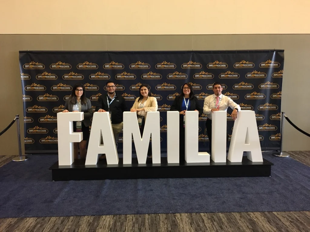 Five people stand behind large white letters spelling "FAMILIA" in front of a branded SHPE backdrop, representing Hispanic Professional Engineers at Valparaiso University.