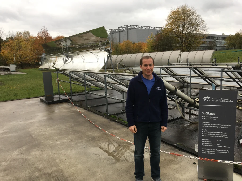 A man stands smiling in front of a solar research installation and an information sign outdoors in Germany, showcasing Professor Venstrom's ongoing research.